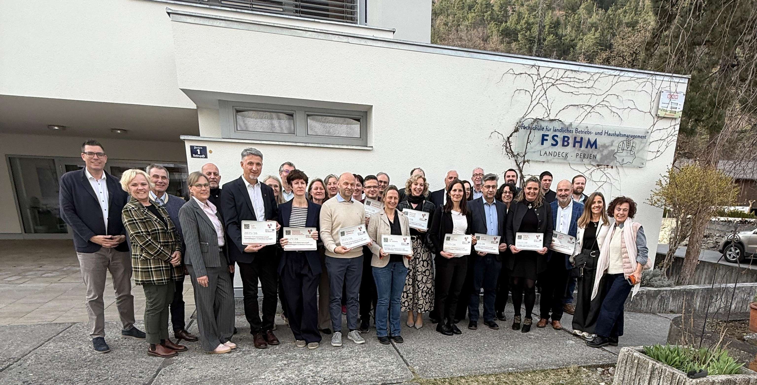 Gruppe von Menschen posiert vor einem Gebäude mit FSBHM-Schild. Einige halten Zertifikate. Hintergrund zeigt Bäume und Berge.