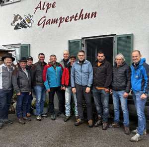 Gruppe von Männern in Outdoorkleidung vor dem Gebäude der Alpe Gamperthun mit Schriftzug und Kuh-Logo an der Fassade.