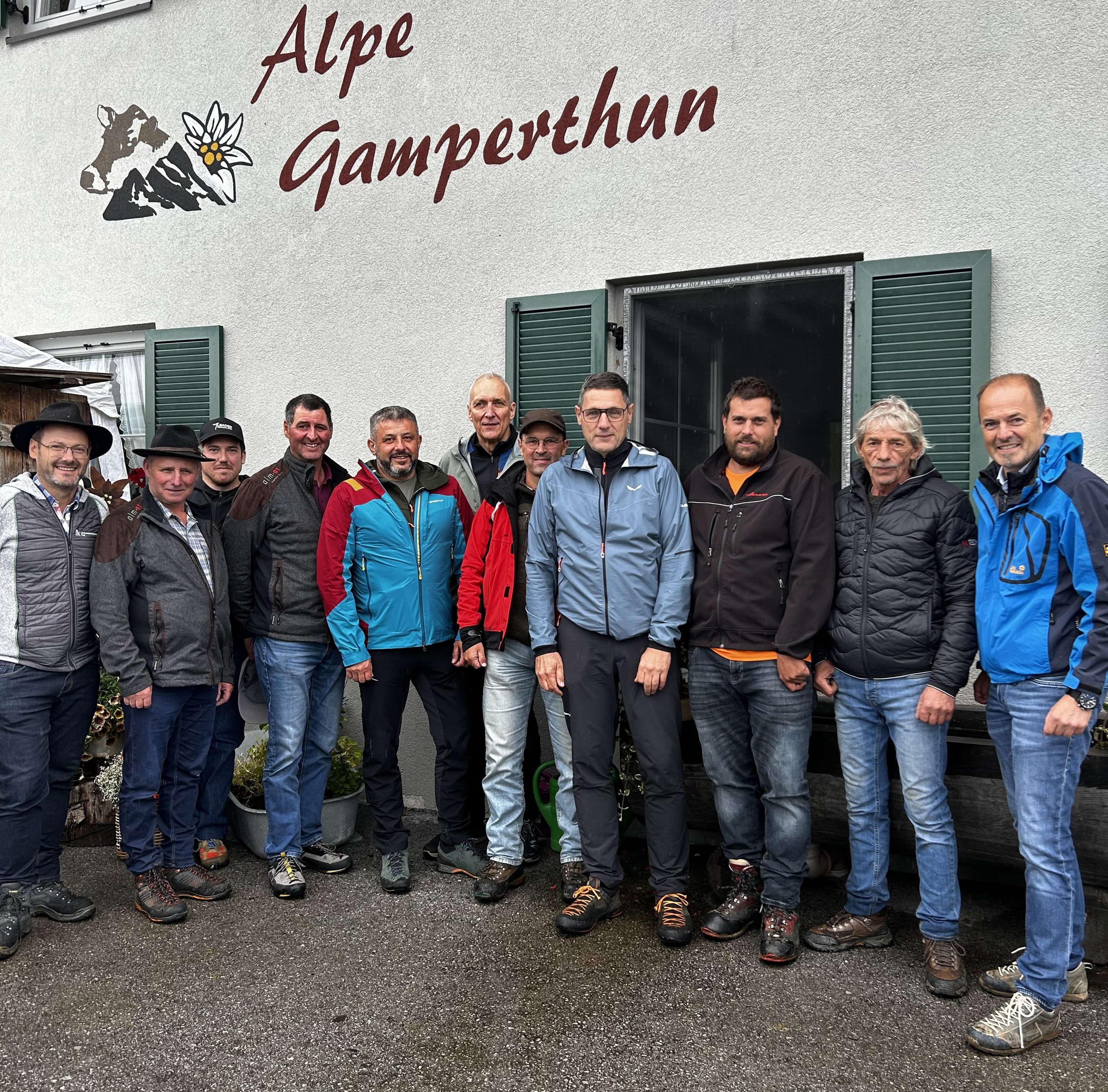 Gruppe von Männern in Outdoorkleidung vor dem Gebäude der Alpe Gamperthun mit Schriftzug und Kuh-Logo an der Fassade.