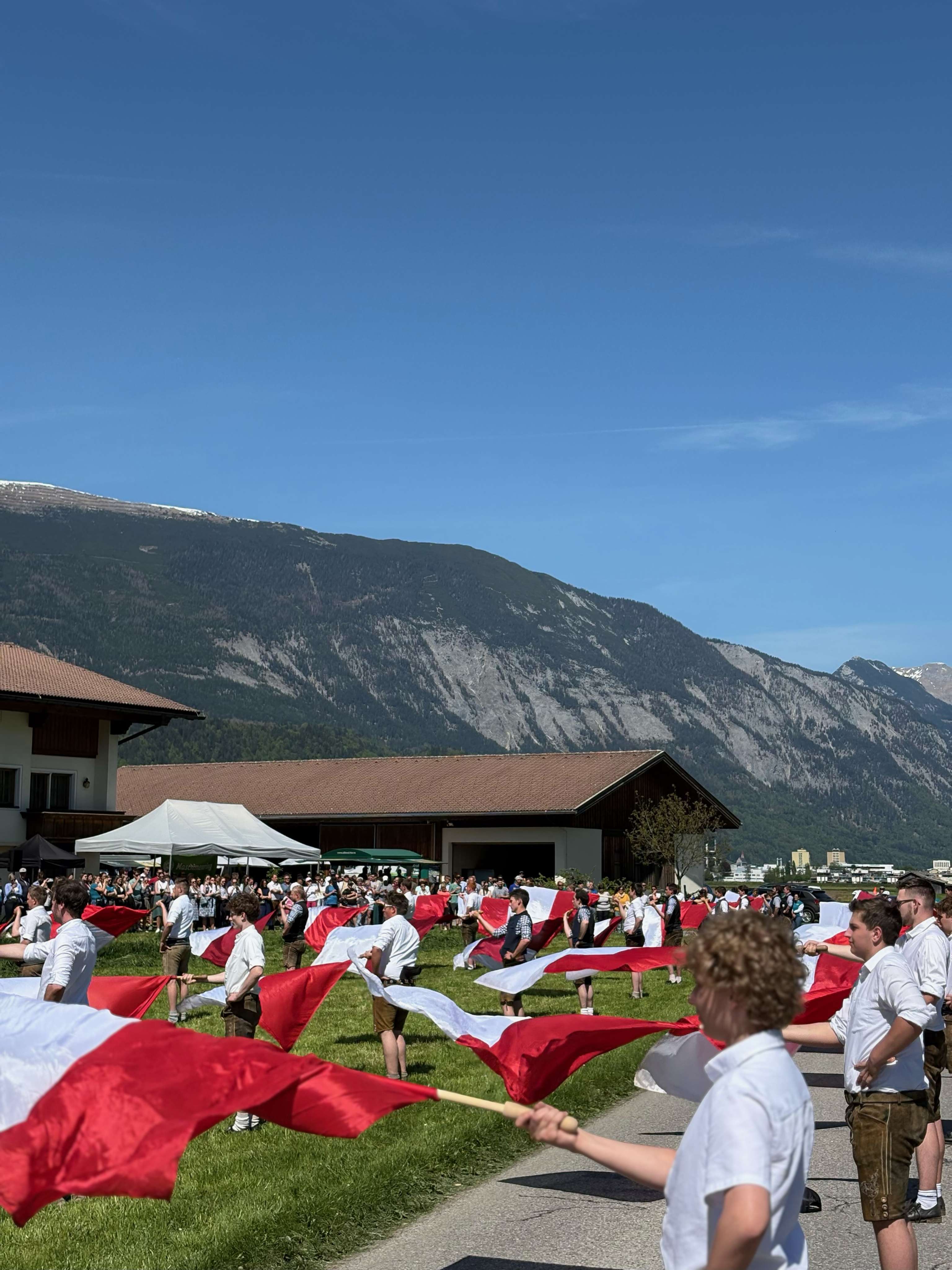Menschen mit rot-weißen Flaggen vor einer Berglandschaft, tragen traditionelle Kleidung, klare Tageslichtszene.