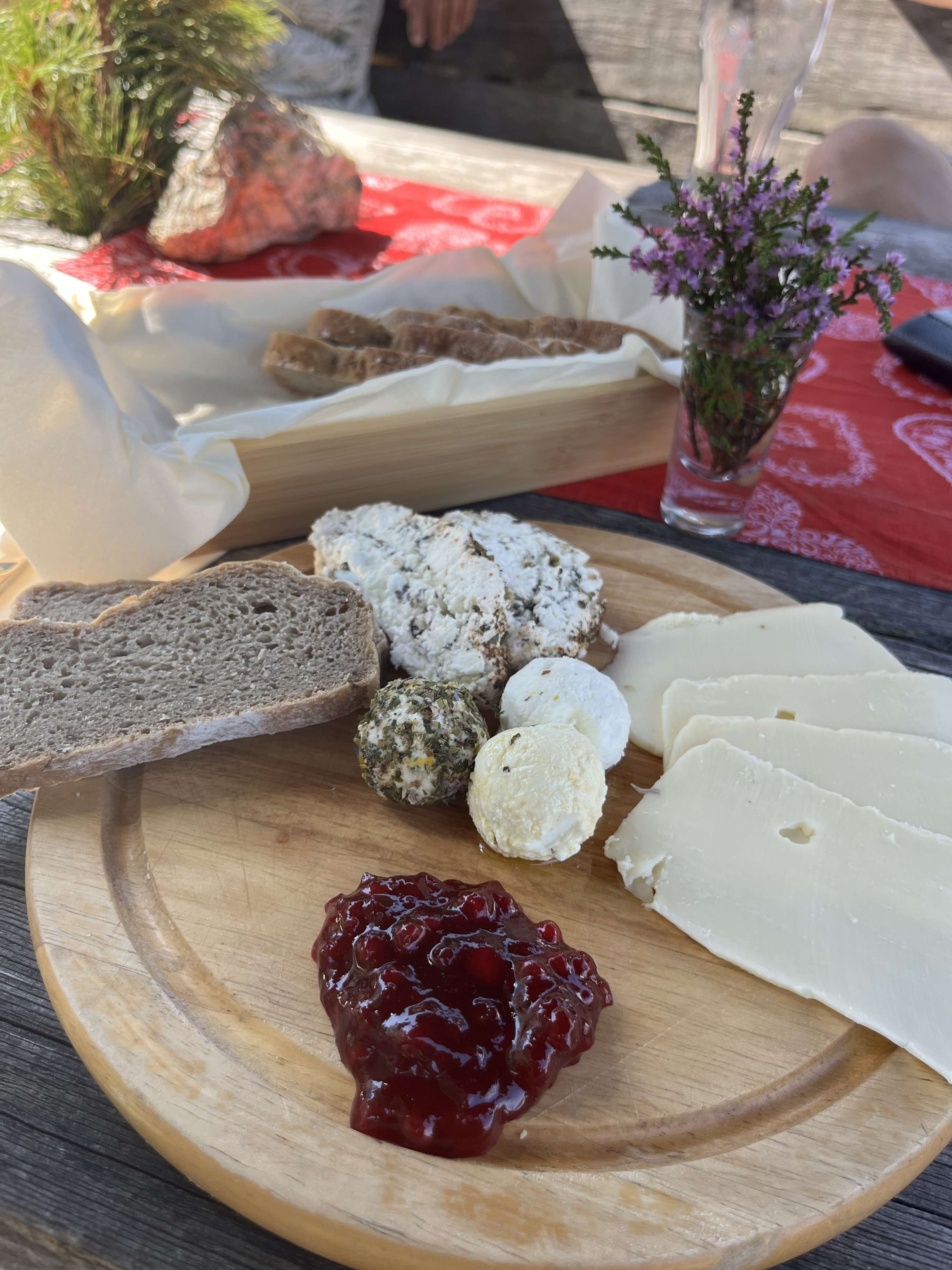 Rustikale Käseplatte mit verschiedenen Käsesorten, Brot und Preiselbeerkonfitüre auf einem Holzbrett, rote Tischdecke mit Heideblumen im Hintergrund.