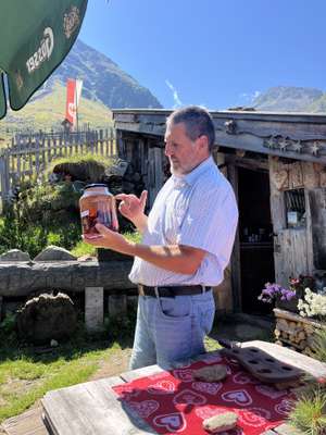 Georg Mair hält ein Einmachglas mit angesetzten Zirbenzapfen vor einer Almhütte in den Alpen bei strahlendem Sonnenschein und Bergen im Hintergrund.