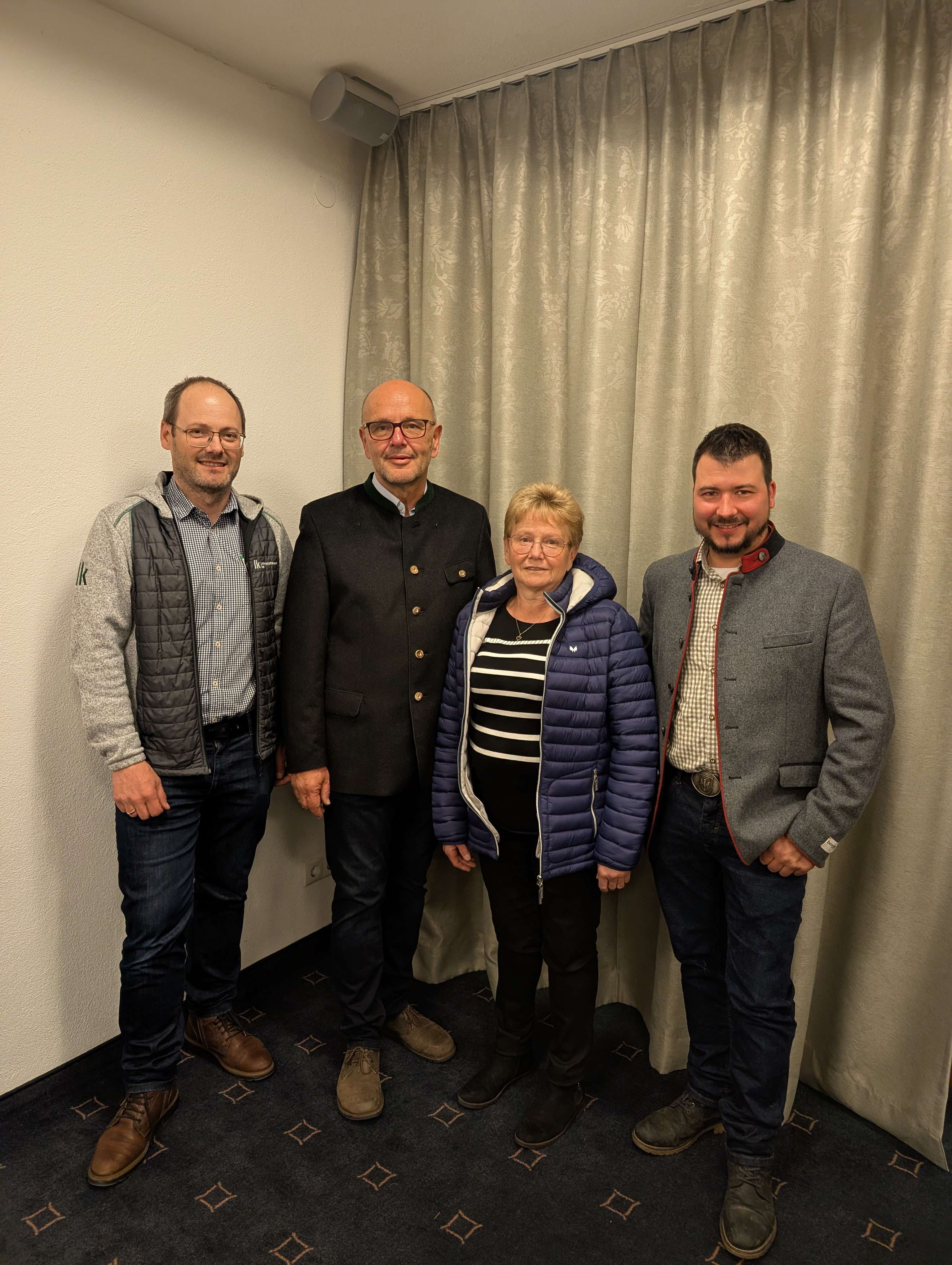 Vier Personen posieren lächelnd in einem Raum vor einem Vorhang. Sie tragen Freizeitkleidung und stehen auf einem Teppich.