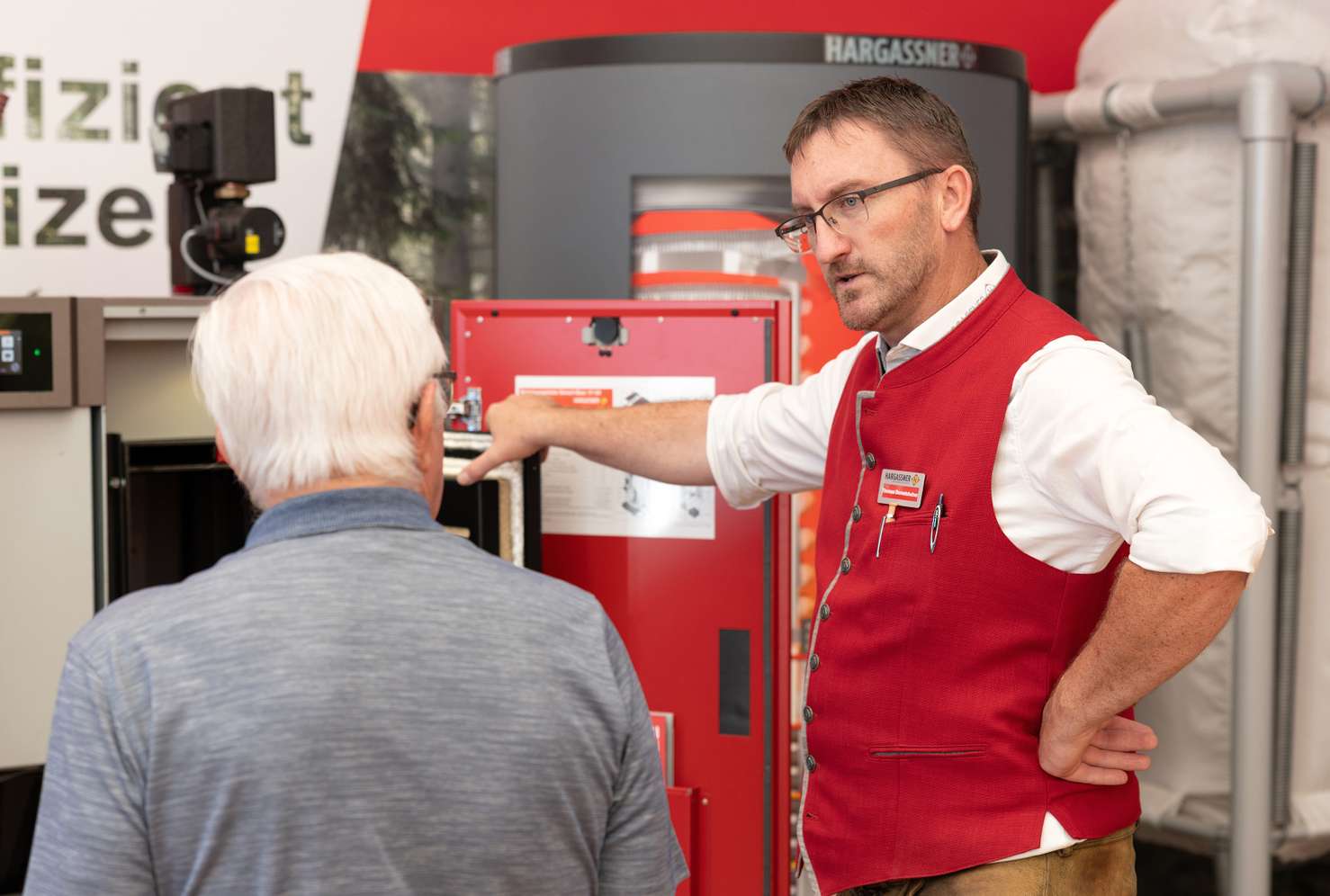 Mann in roter Weste erklärt einem älteren Mann ein technisches Gerät auf einer Messe mit dem Logo "Hargassner" im Hintergrund.