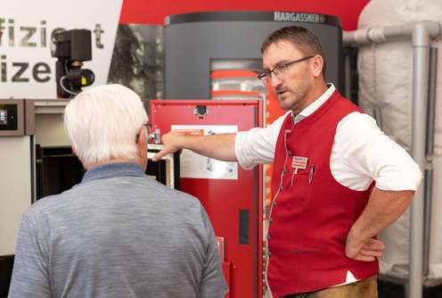 Mann in roter Weste erklärt einem älteren Mann ein technisches Gerät auf einer Messe mit dem Logo "Hargassner" im Hintergrund.