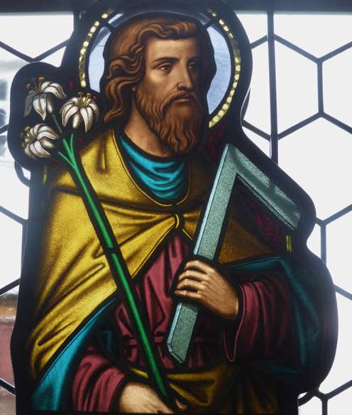 Stained glass of a bearded man holding white lilies and a carpenter's square, with a halo, set against a hexagonal patterned window.