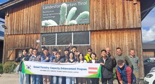 A group of people holding a banner for the Global Forestry Capacity Enhancement Program, standing in front of a wooden building in Tirol.