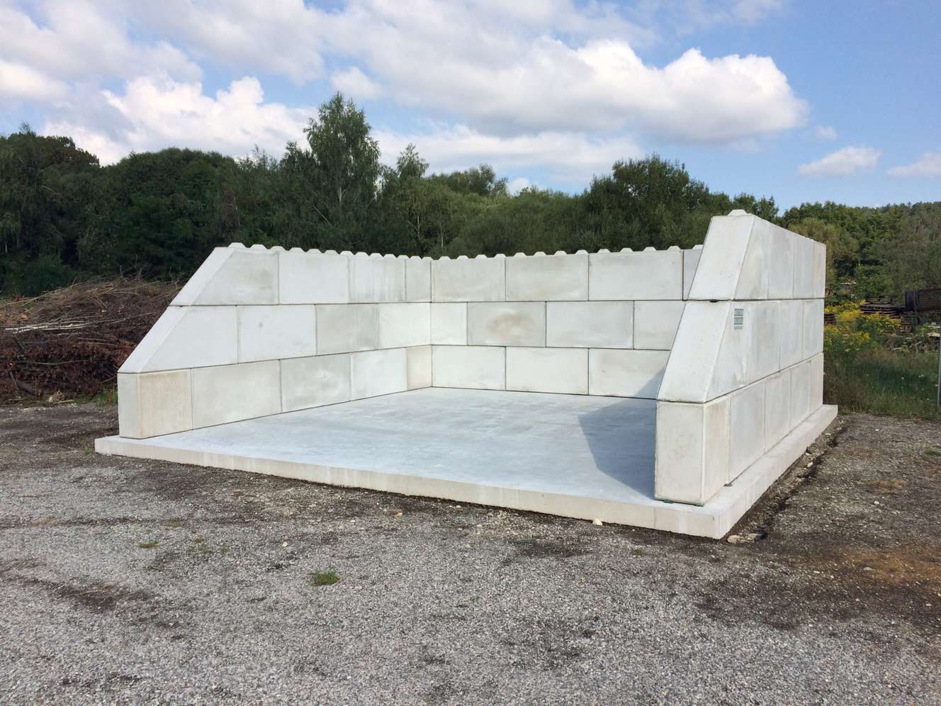 Betonfertigteil-Konstruktion mit U-förmiger Wandstruktur und Bodenplatte auf Kiesgelände, umgeben von Bäumen unter bewölktem Himmel.