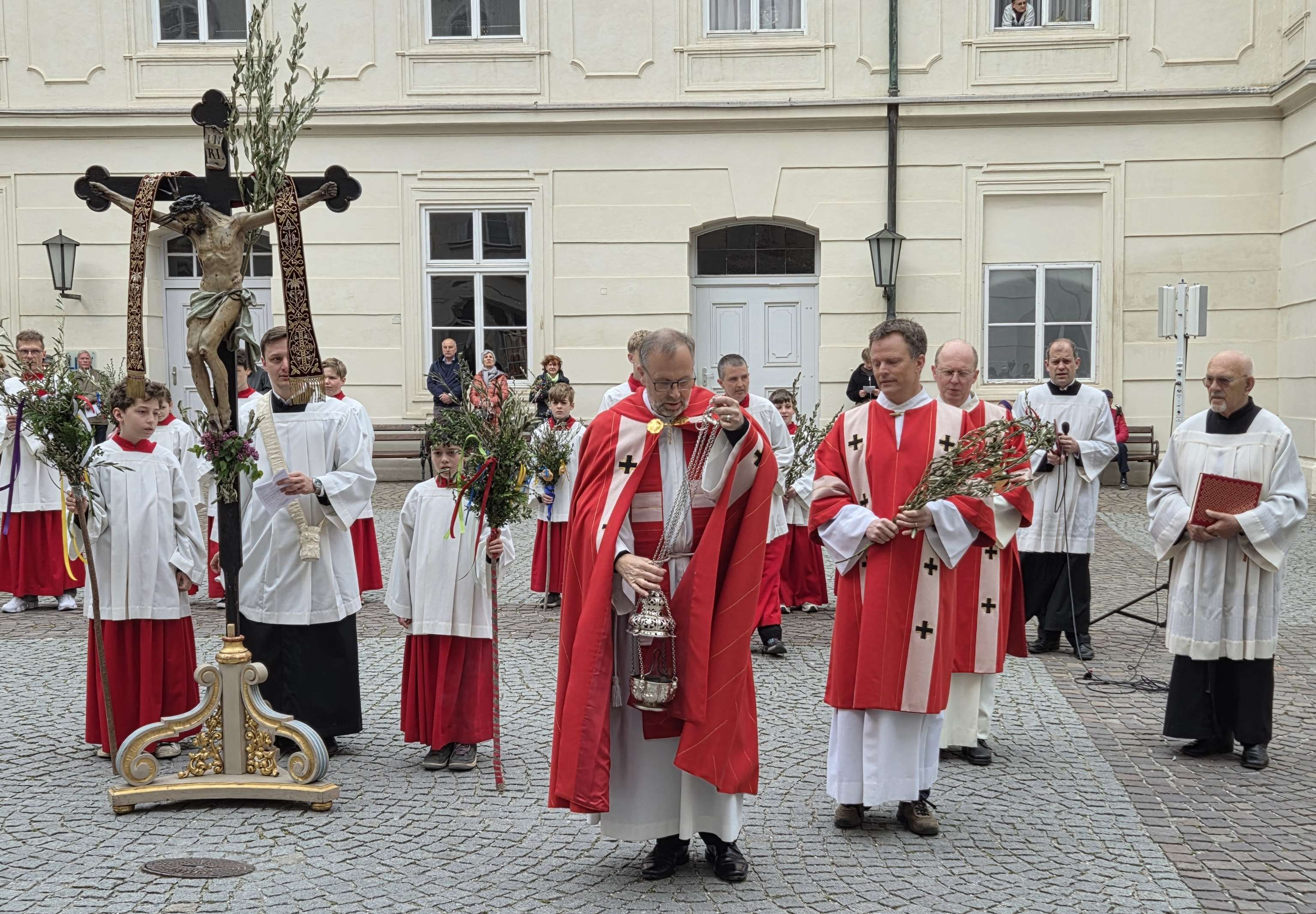 Priester und Ministranten in Rot und Weiß tragen Kruzifix und Zweige bei einer religiösen Prozession auf einem gepflasterten Platz.