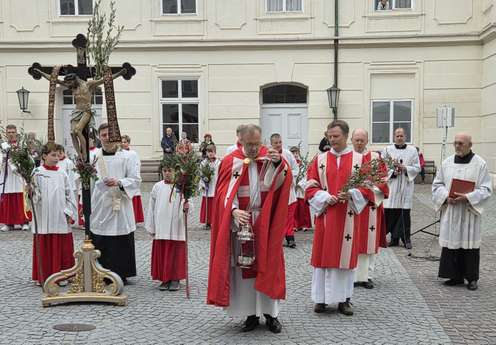 Priester und Ministranten in Rot und Weiß tragen Kruzifix und Zweige bei einer religiösen Prozession auf einem gepflasterten Platz.