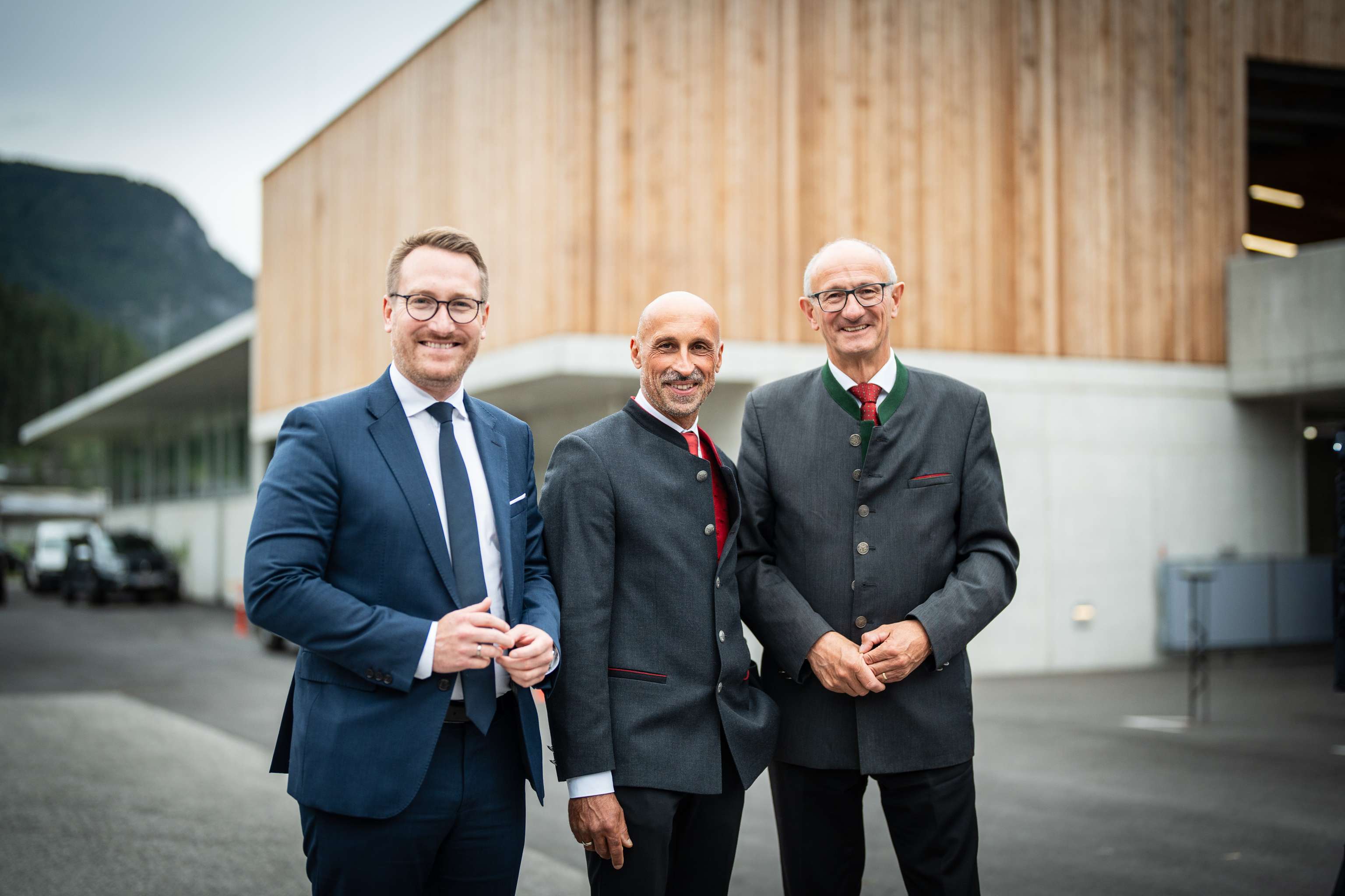 Drei lächelnde Männer in Anzügen und Trachtenjacken vor einem modernen Holzgebäude mit Bergkulisse.