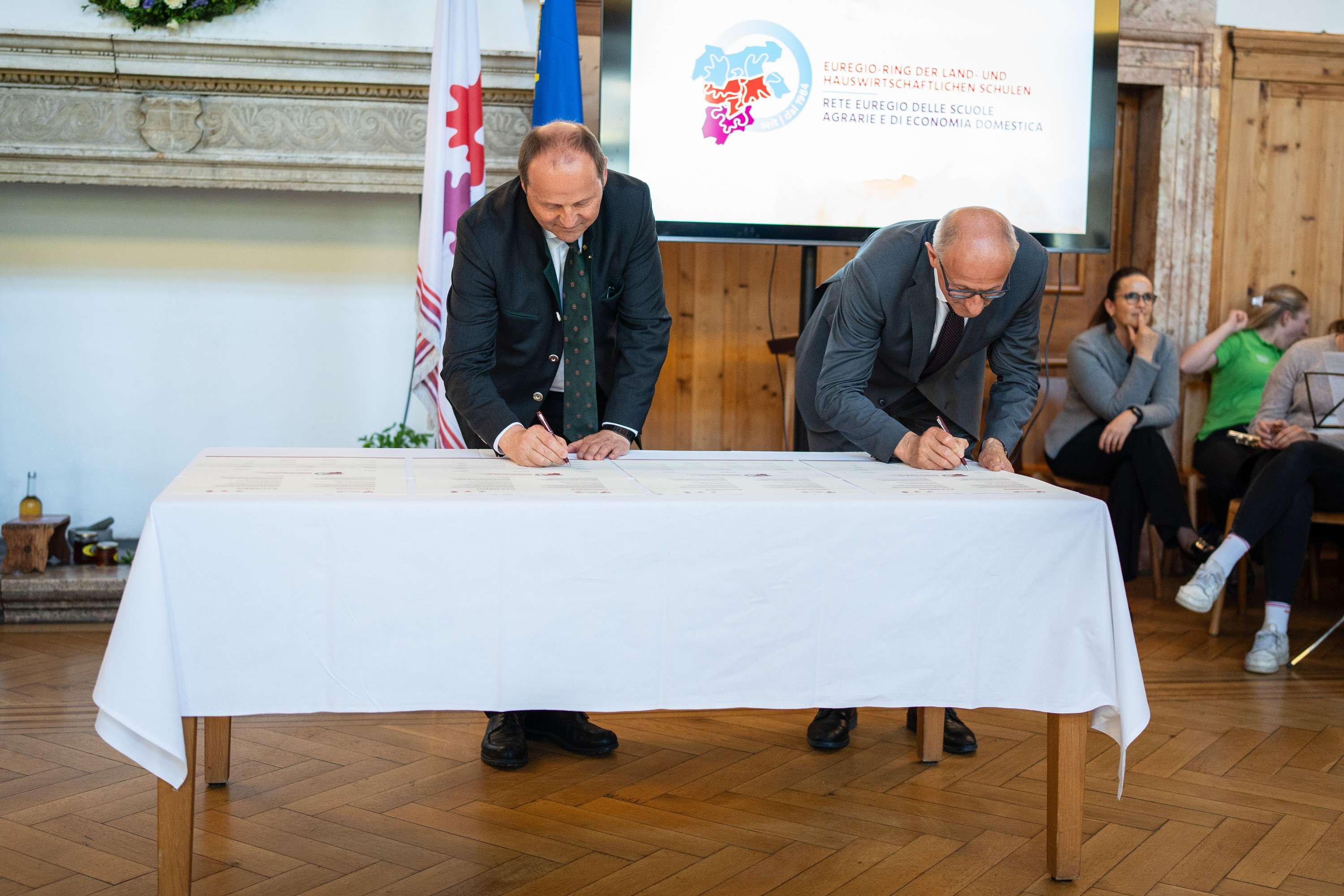 Zwei Männer unterschreiben Dokumente auf einem Tisch. Im Hintergrund sitzt eine Gruppe von Menschen. Ein Bildschirm zeigt ein Logo.