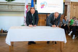 Zwei Männer unterschreiben Dokumente auf einem Tisch. Im Hintergrund sitzt eine Gruppe von Menschen. Ein Bildschirm zeigt ein Logo.