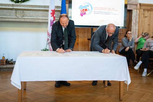 Zwei Männer unterschreiben Dokumente auf einem Tisch. Im Hintergrund sitzt eine Gruppe von Menschen. Ein Bildschirm zeigt ein Logo.