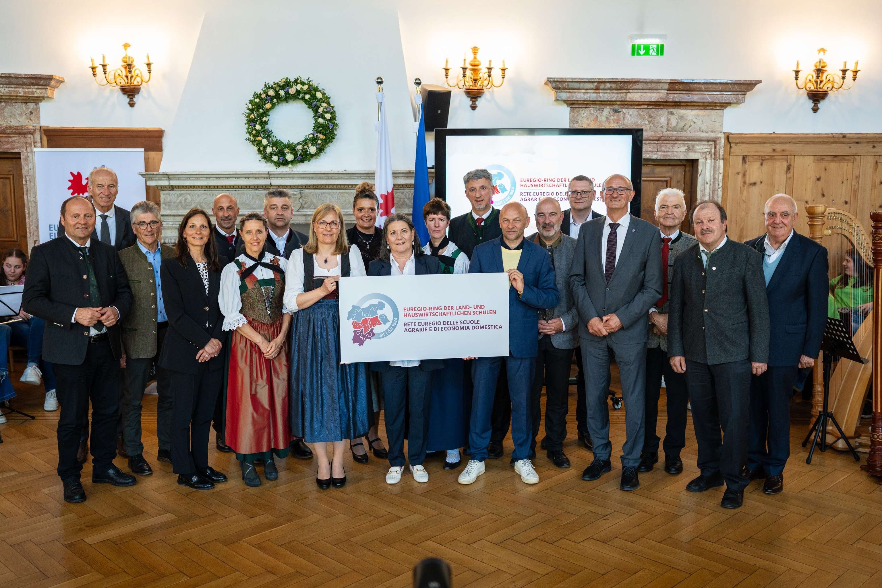 Gruppe von Menschen in traditioneller und formeller Kleidung hält ein Banner mit Text in einem festlichen Raum mit Holzfußboden.