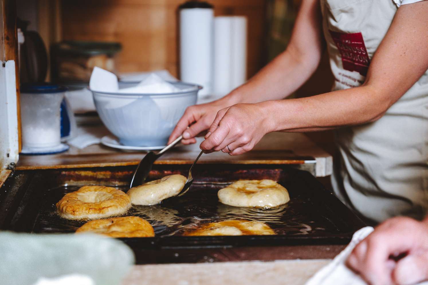 Frau in Schürze wendet goldbraune Hefekrapfen in einer Pfanne mit heißem Öl mithilfe einer Zange.