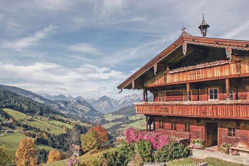 Traditionelles Tiroler Bauernhof mit Balkon und rosa Blumen, mit Blick auf ein grünes Alpental und Berggipfel unter blauem Himmel.