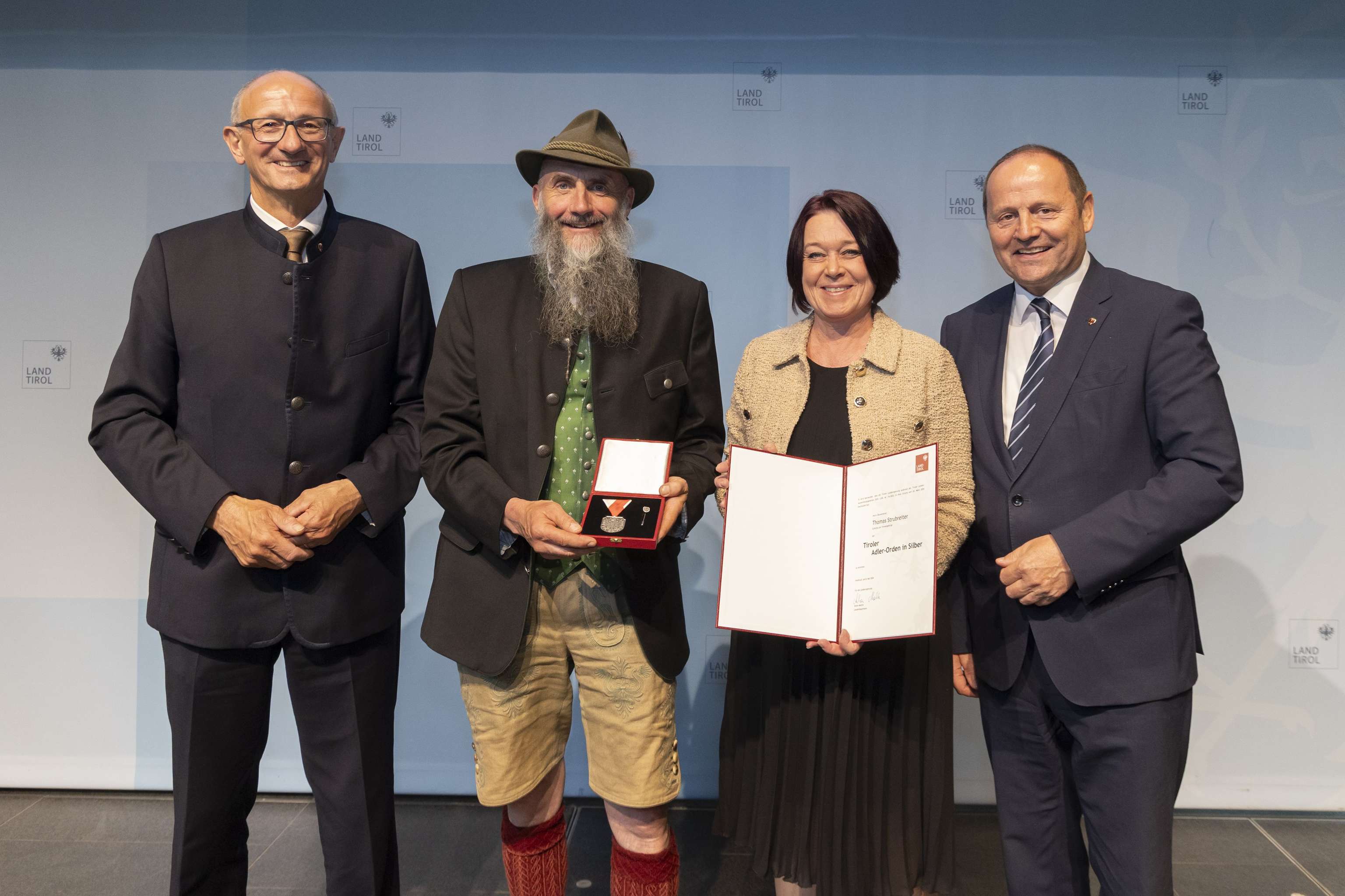 Vier Personen bei einer Preisverleihung in Tirol; eine Frau hält eine Urkunde, ein Mann eine Medaille. Hintergrund: "Land Tirol"-Logo.