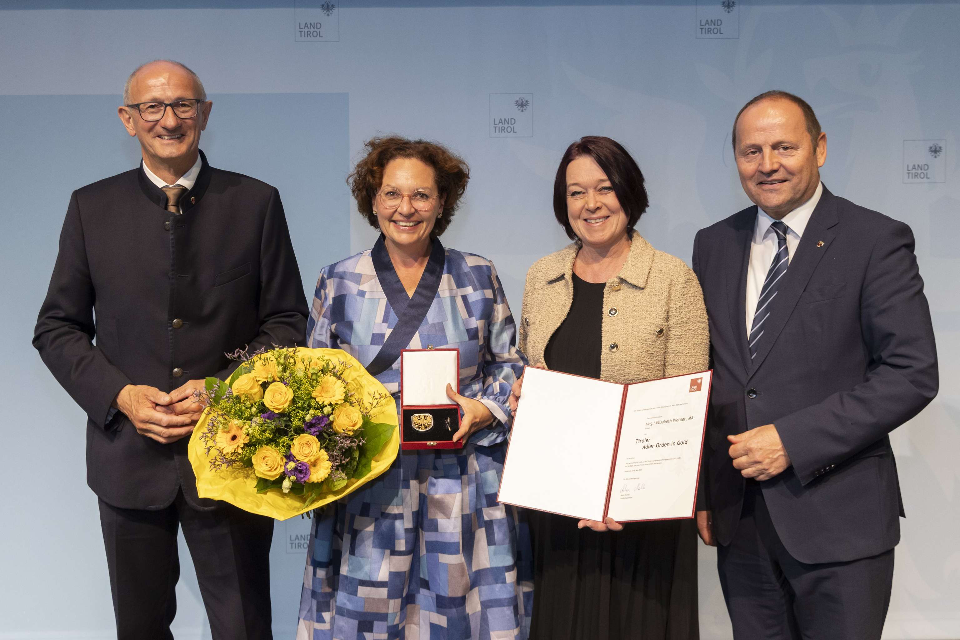 Vier Personen bei einer Auszeichnung, eine Frau hält Blumen, eine andere eine Urkunde. Hintergrund zeigt "Land Tirol"-Symbole.