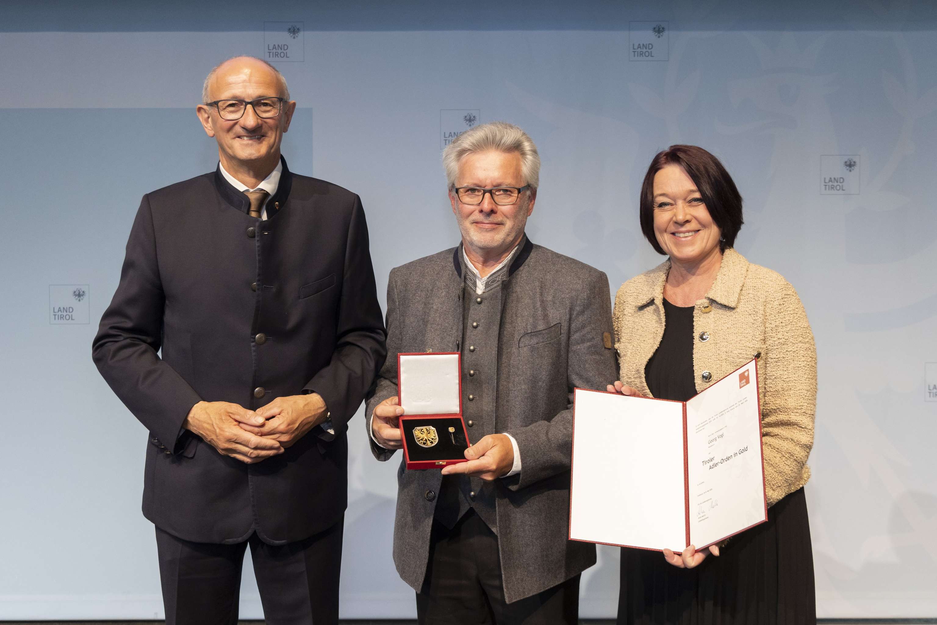 Drei Personen bei einer Preisverleihung, mittlere Person hält eine Medaille, rechte Person eine Urkunde. Hintergrund: Logo "Land Tirol".