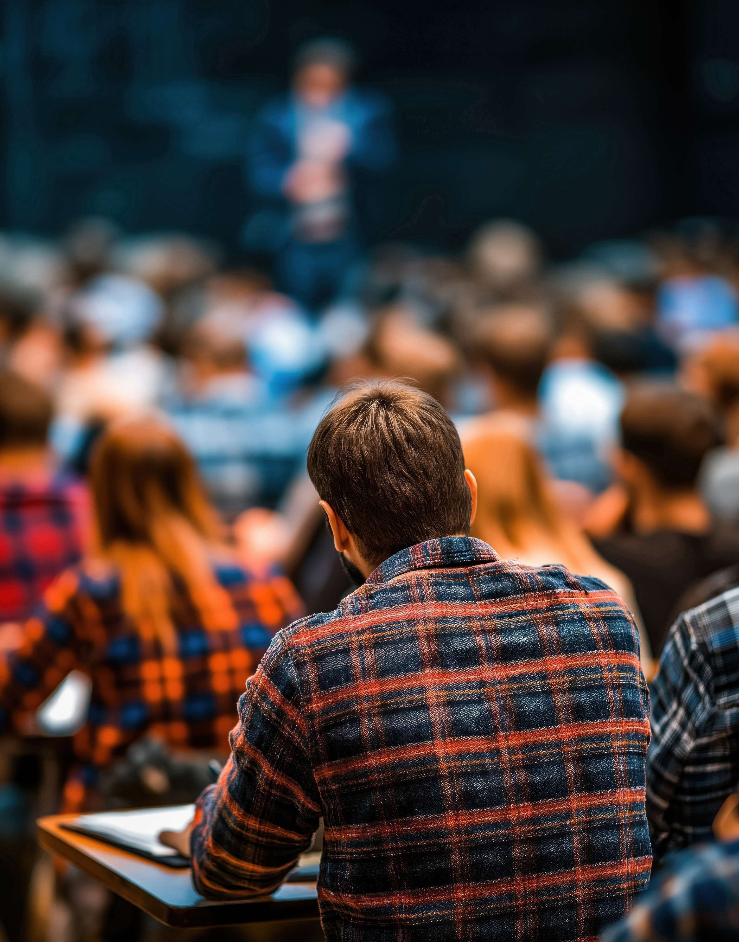 Student im karierten Hemd von hinten in einem vollen Hörsaal, im Hintergrund ein verschwommener Dozent am Podium.