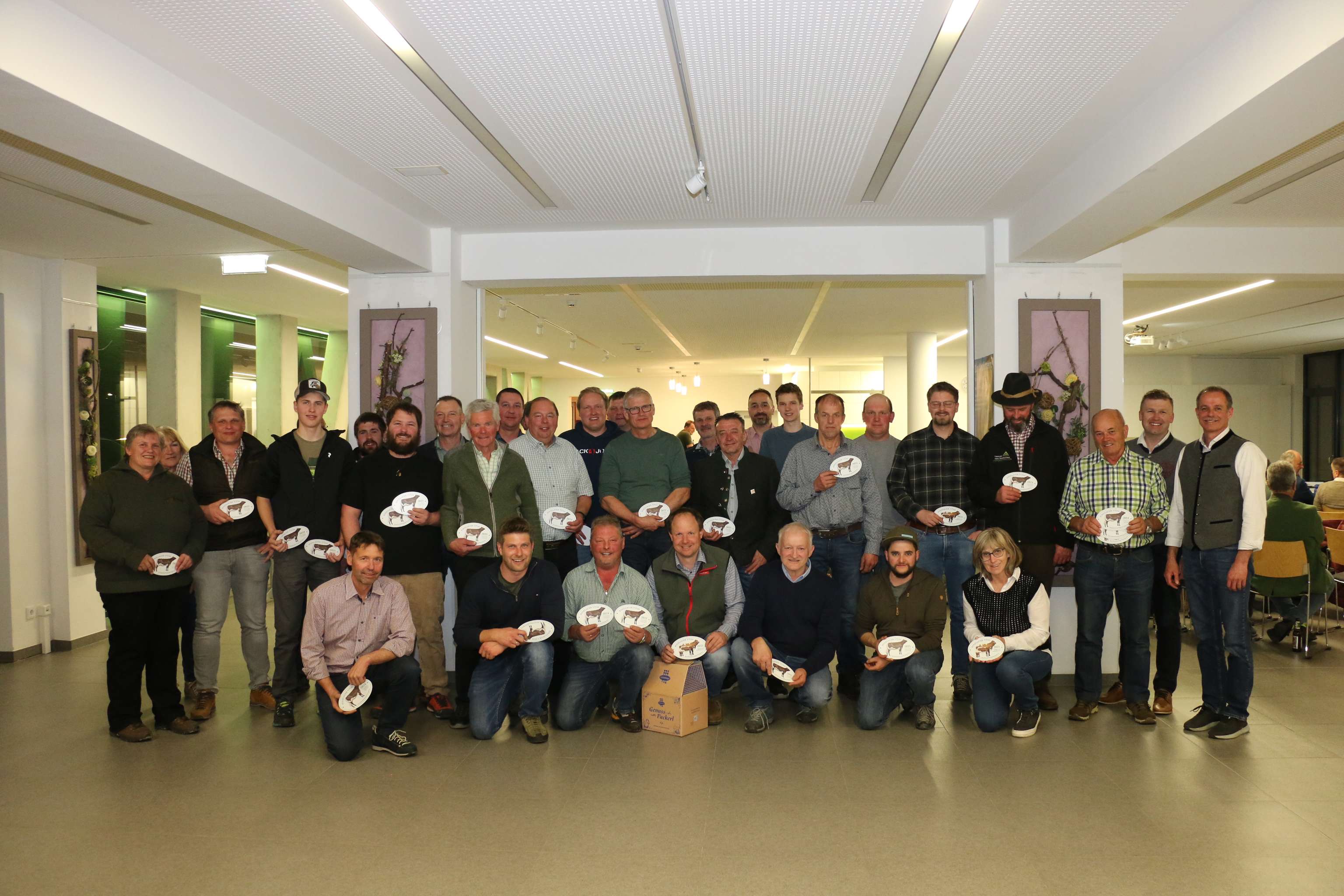 Eine Gruppe von Menschen steht in einem Innenraum, viele halten Teller. Es sind insgesamt etwa 28 Personen, darunter einige in grüner Kleidung.