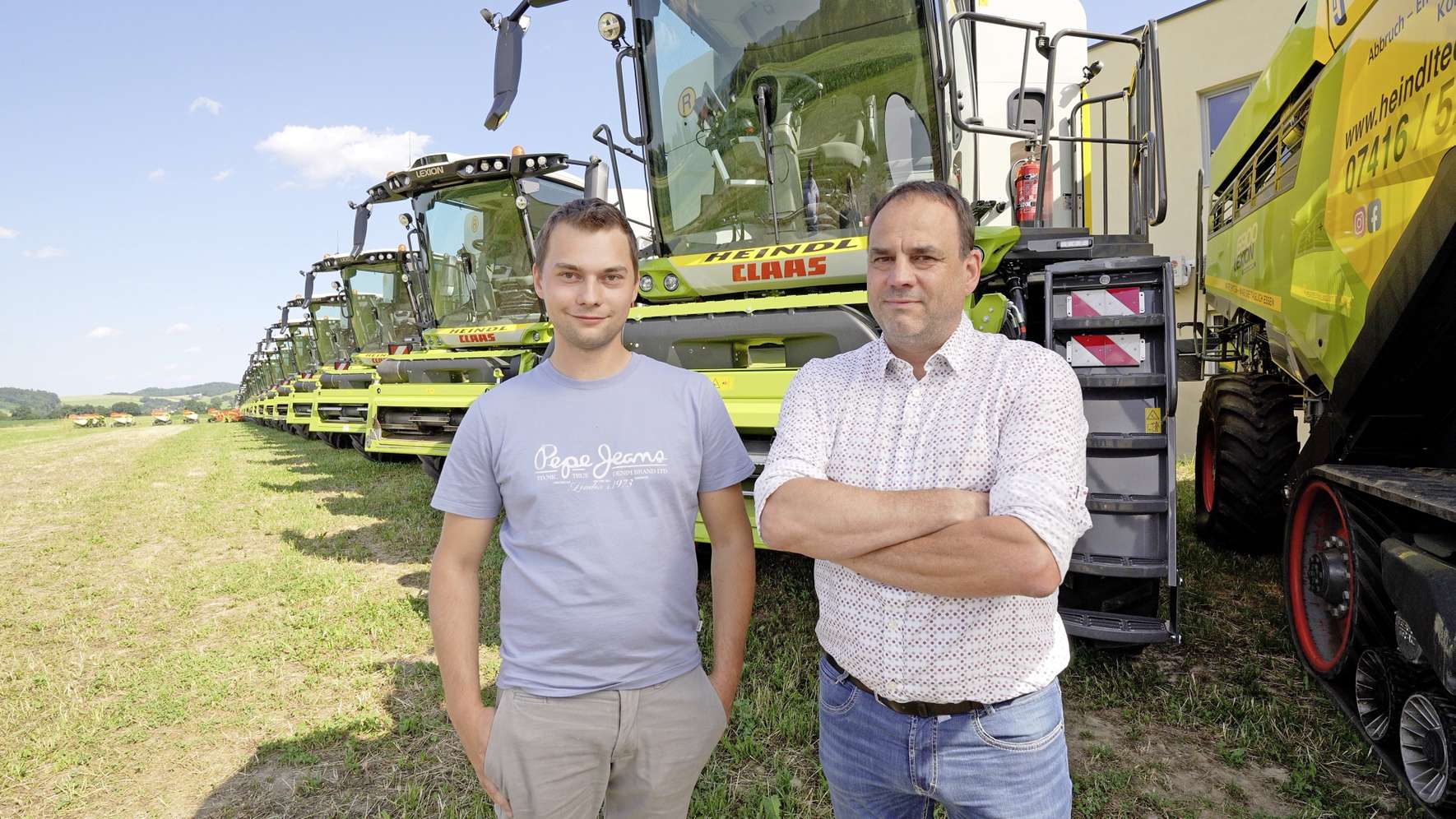 Two men stand in front of a row of large green agricultural machines on a sunny day, with fields and trees in the background.