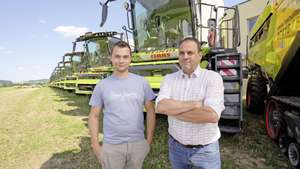 Two men stand in front of a row of large green agricultural machines on a sunny day, with fields and trees in the background.