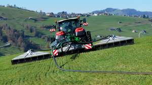 A tractor with large attachments operates on a sloped green field, with rolling hills and scattered houses in the background.