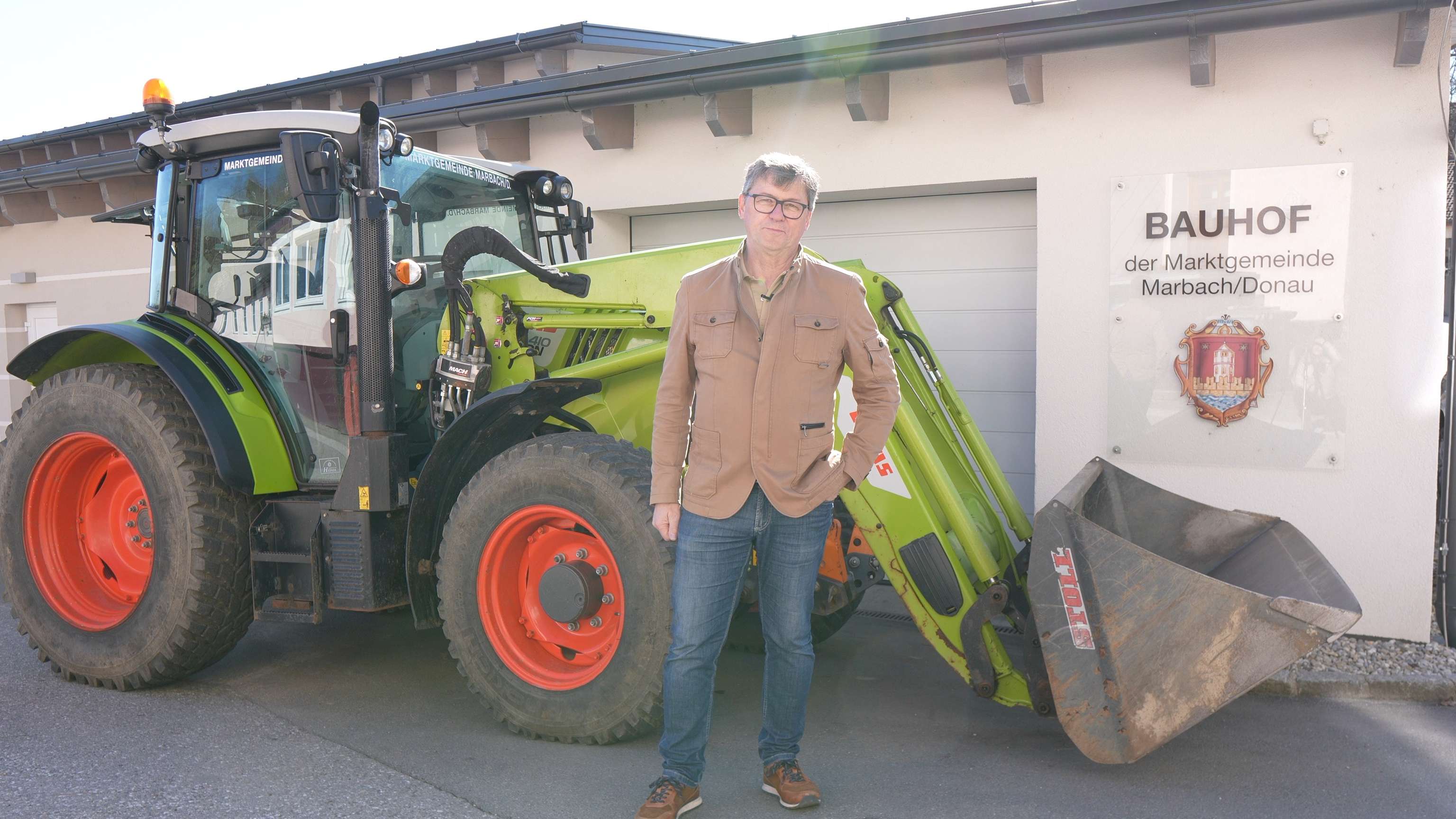 Man standing in front of a green tractor with a scoop, beside a building labeled "BAUHOF der Marktgemeinde Marbach/Donau."