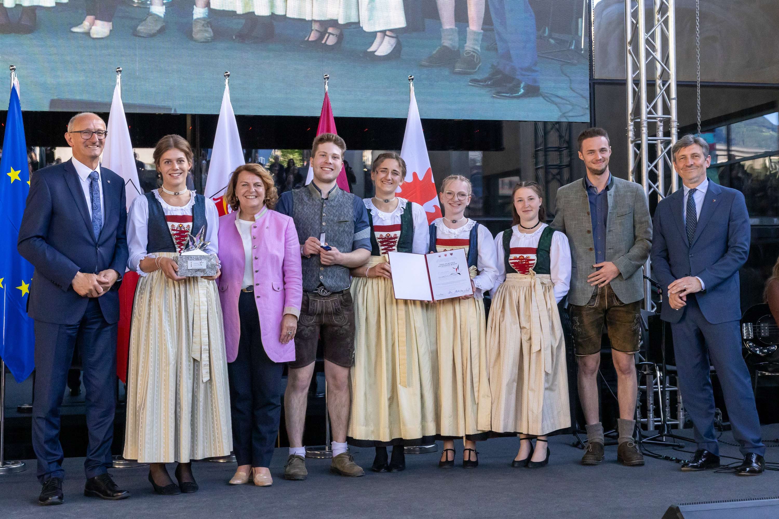 Eine Gruppe von Personen in traditioneller Kleidung steht auf einer Bühne, einige halten Trophäen und Zertifikate, mit Flaggen im Hintergrund.