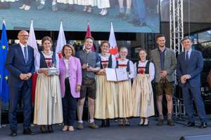 Eine Gruppe von Personen in traditioneller Kleidung steht auf einer Bühne, einige halten Trophäen und Zertifikate, mit Flaggen im Hintergrund.