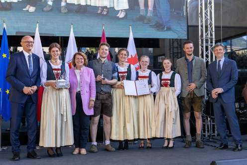 Eine Gruppe von Personen in traditioneller Kleidung steht auf einer Bühne, einige halten Trophäen und Zertifikate, mit Flaggen im Hintergrund.