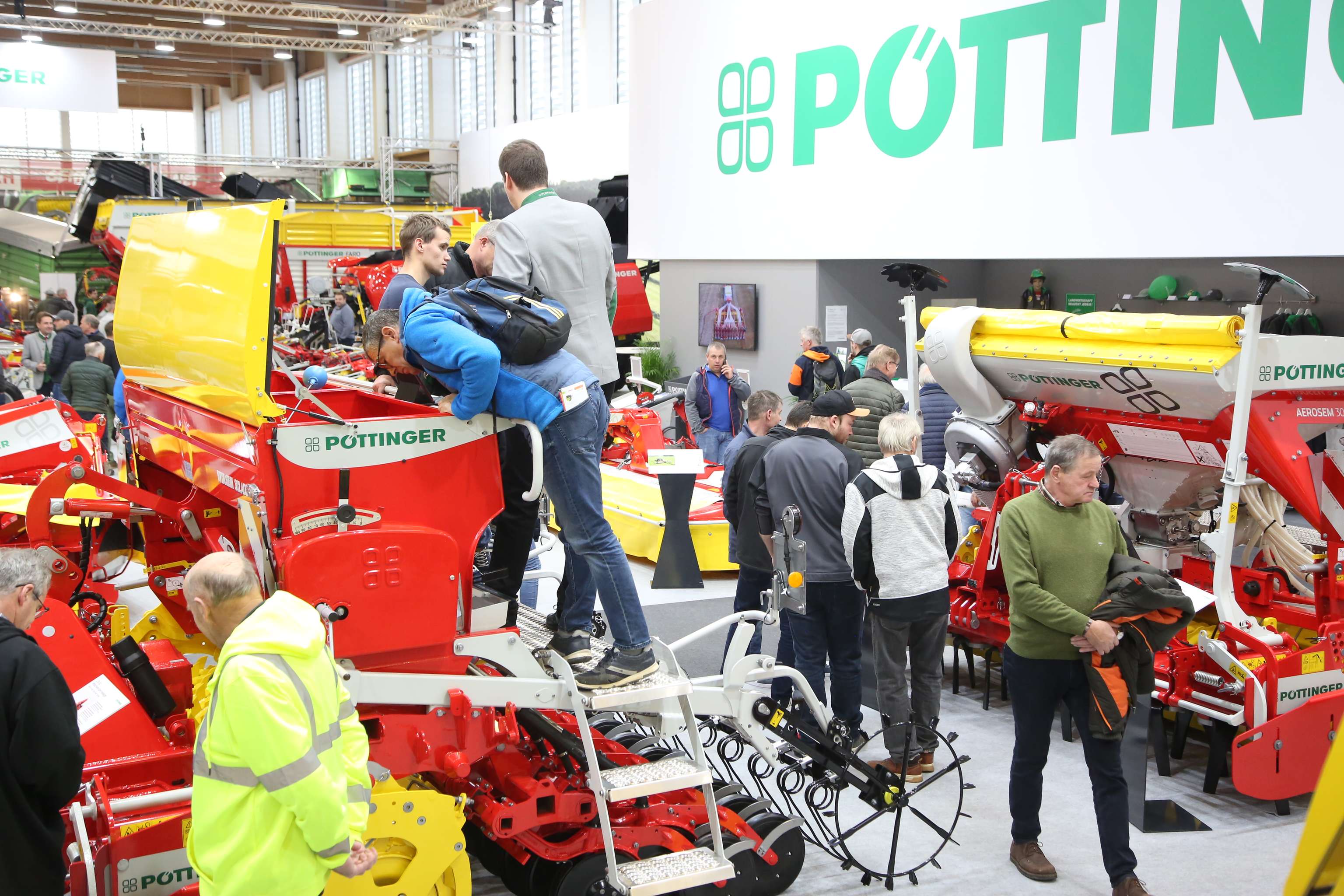 Messestand von Pöttinger mit roten und gelben Landmaschinen, umgeben von zahlreichen Besuchern in einer Ausstellungshalle.