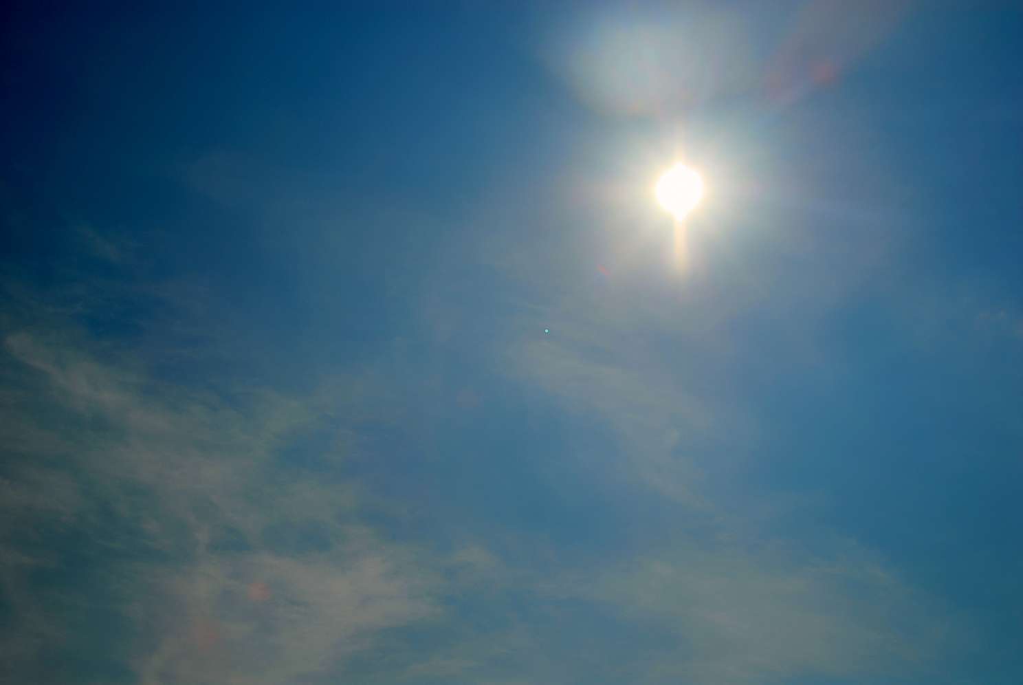 Strahlende Sonne mit Linsenreflexion am blauen Himmel, vereinzelte Wolken im Vordergrund.