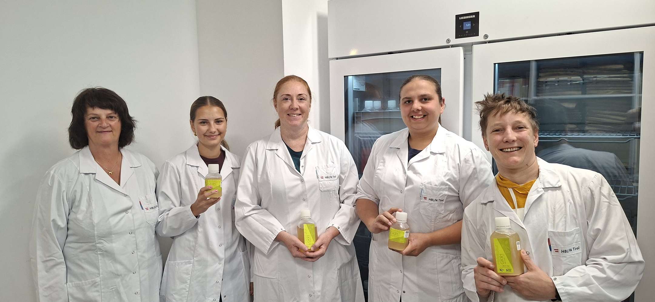 Fünf Frauen in weißen Laborkitteln der HBLFA Tirol stehen im Labor und halten gelbe Probenflaschen in den Händen.