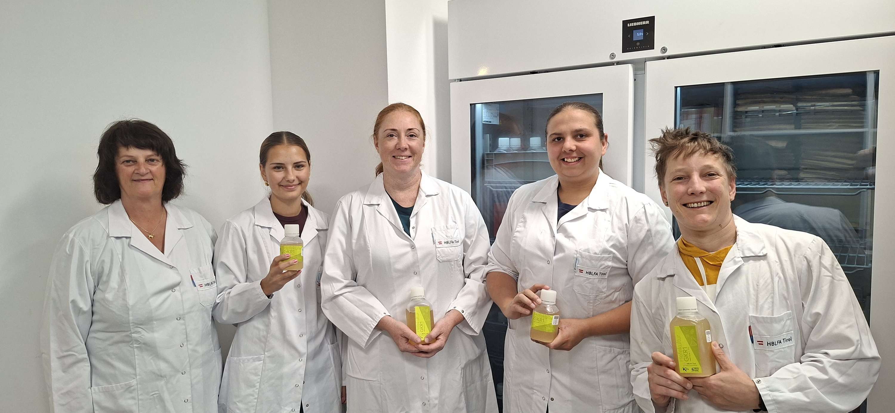 Fünf Frauen in weißen Laborkitteln der HBLFA Tirol stehen im Labor und halten gelbe Probenflaschen in den Händen.