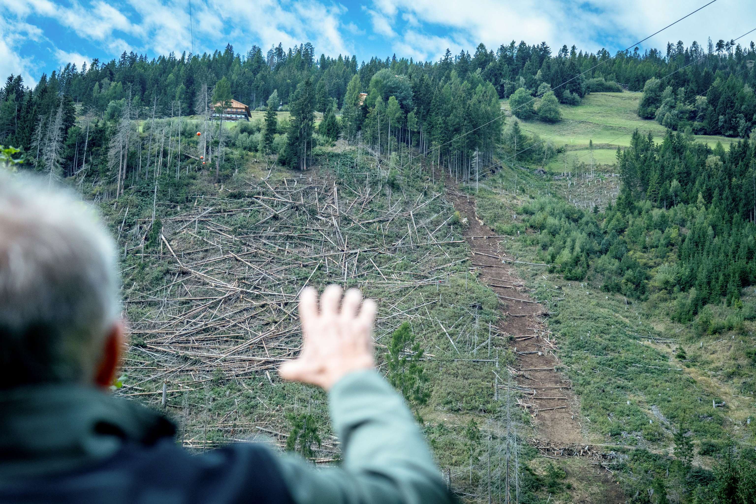 Mann zeigt auf einen Berghang mit massivem Windwurfschaden, zahlreiche umgestürzte Bäume, im Hintergrund ein Haus und dichter Wald.