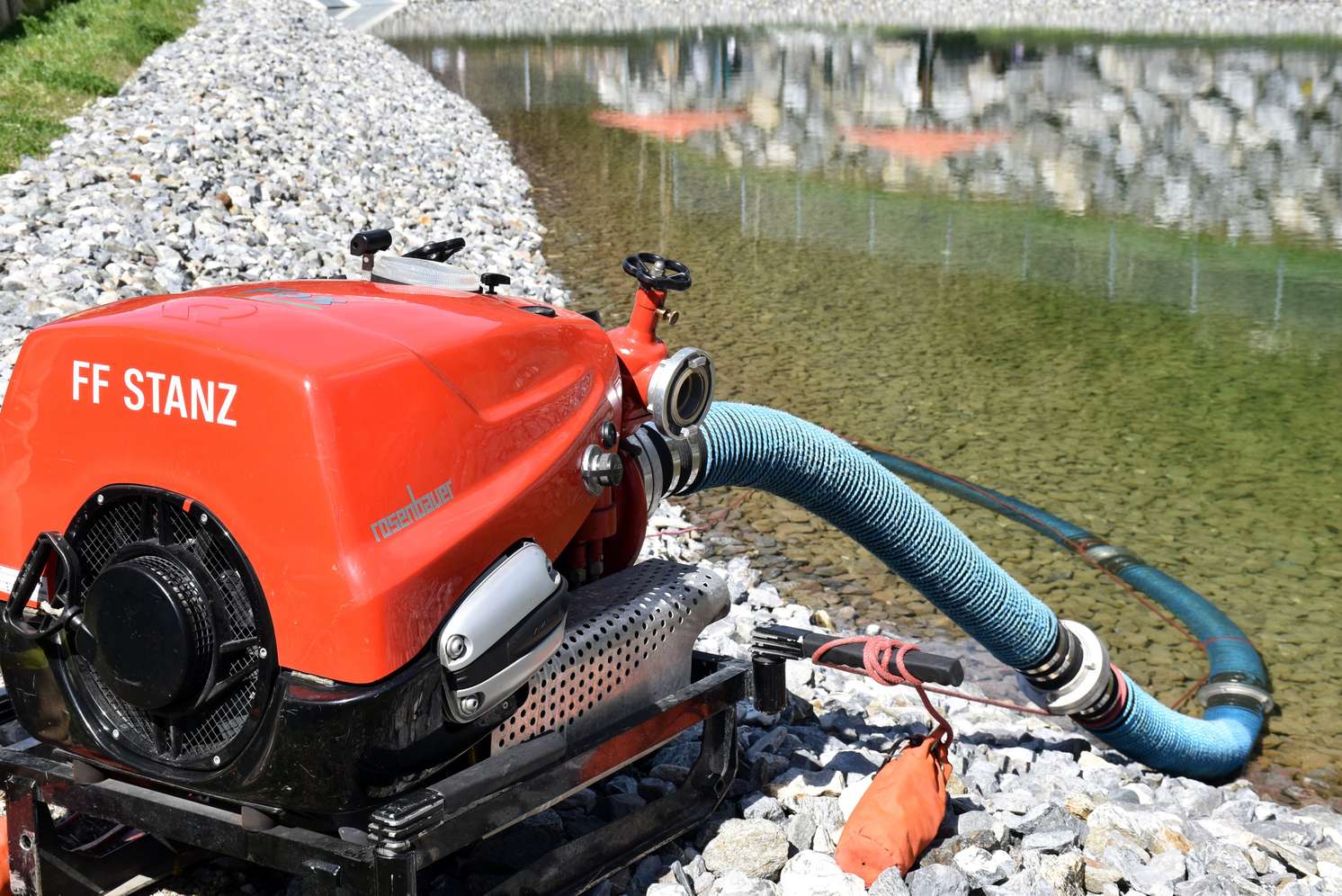 Rote Wasserpumpe von "FF Stanz" am Ufer eines Teichs, mit blauem Schlauch, der ins Wasser führt, auf einem steinigen Untergrund.
