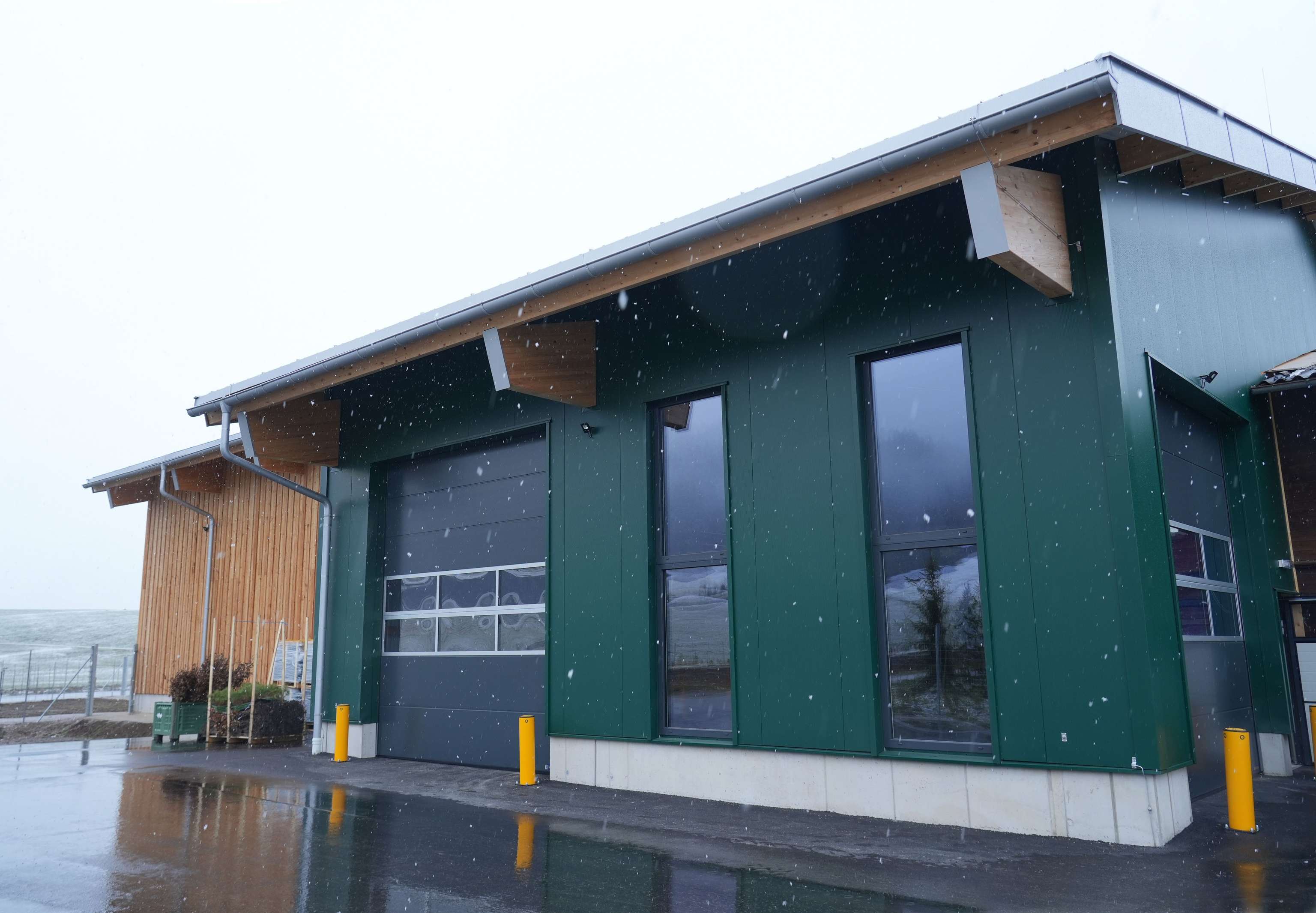 Grünes modernes Gebäude mit großen Fenstern, schrägem Dach und Holzelementen, auf nassem Boden bei leichtem Schneefall.