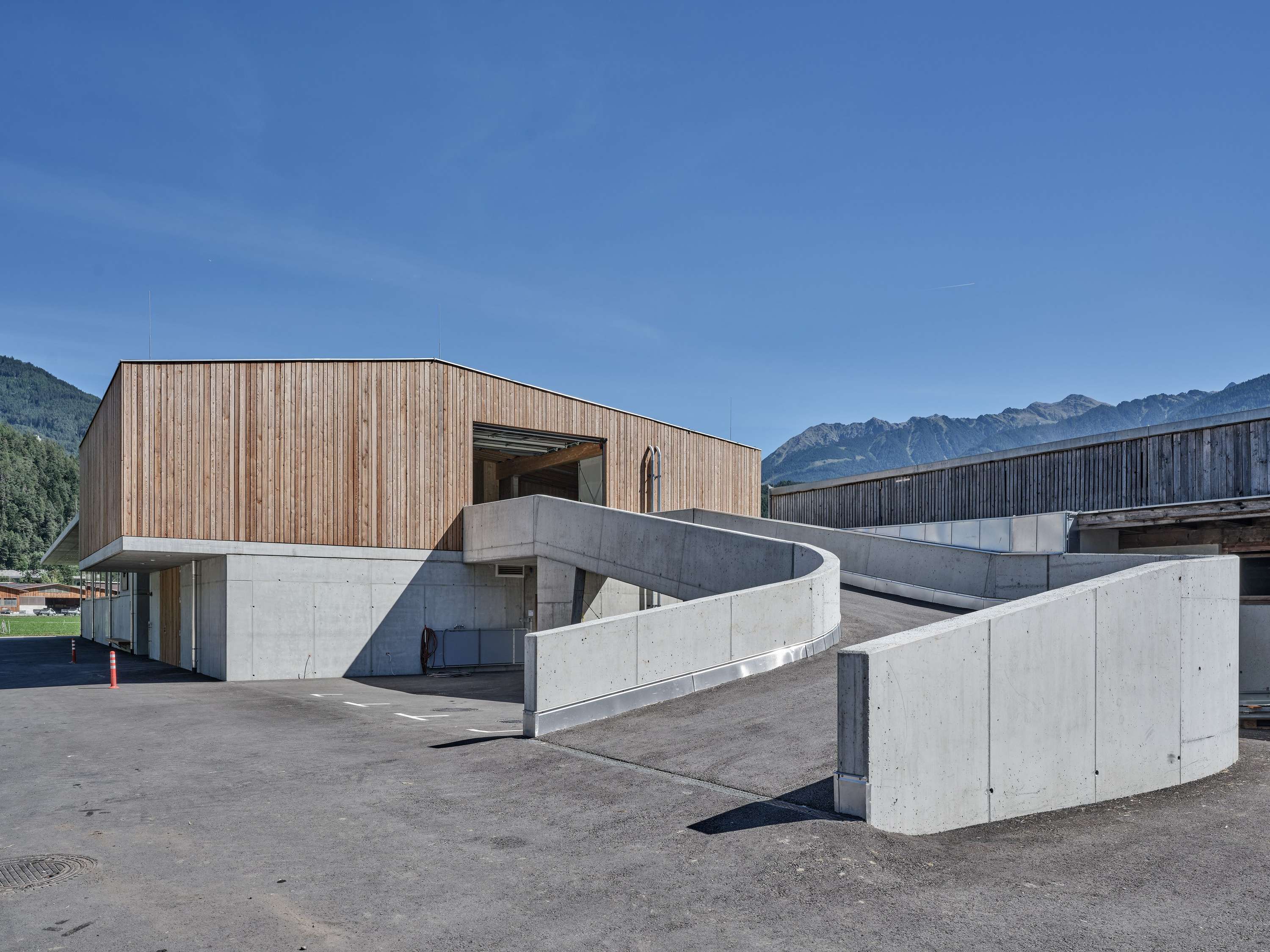 Modernes Gebäude mit Holzfassade und geschwungenen Betonwänden vor Alpenkulisse unter blauem Himmel.