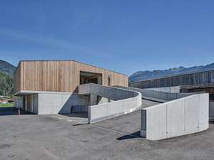Modernes Gebäude mit Holzfassade und geschwungenen Betonwänden vor Alpenkulisse unter blauem Himmel.