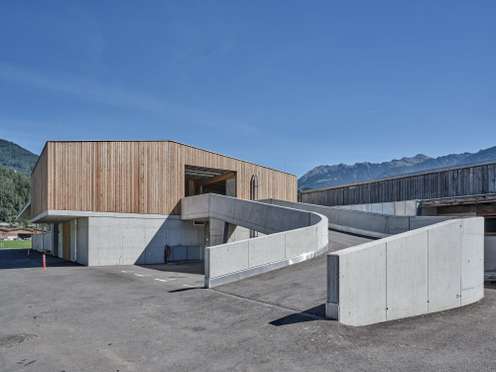 Modernes Gebäude mit Holzfassade und geschwungenen Betonwänden vor Alpenkulisse unter blauem Himmel.