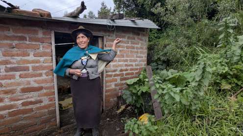 Indigene Frau mit Hut und blauem Umhang steht vor einem Backsteingebäude auf dem Land und zeigt auf das Dach.