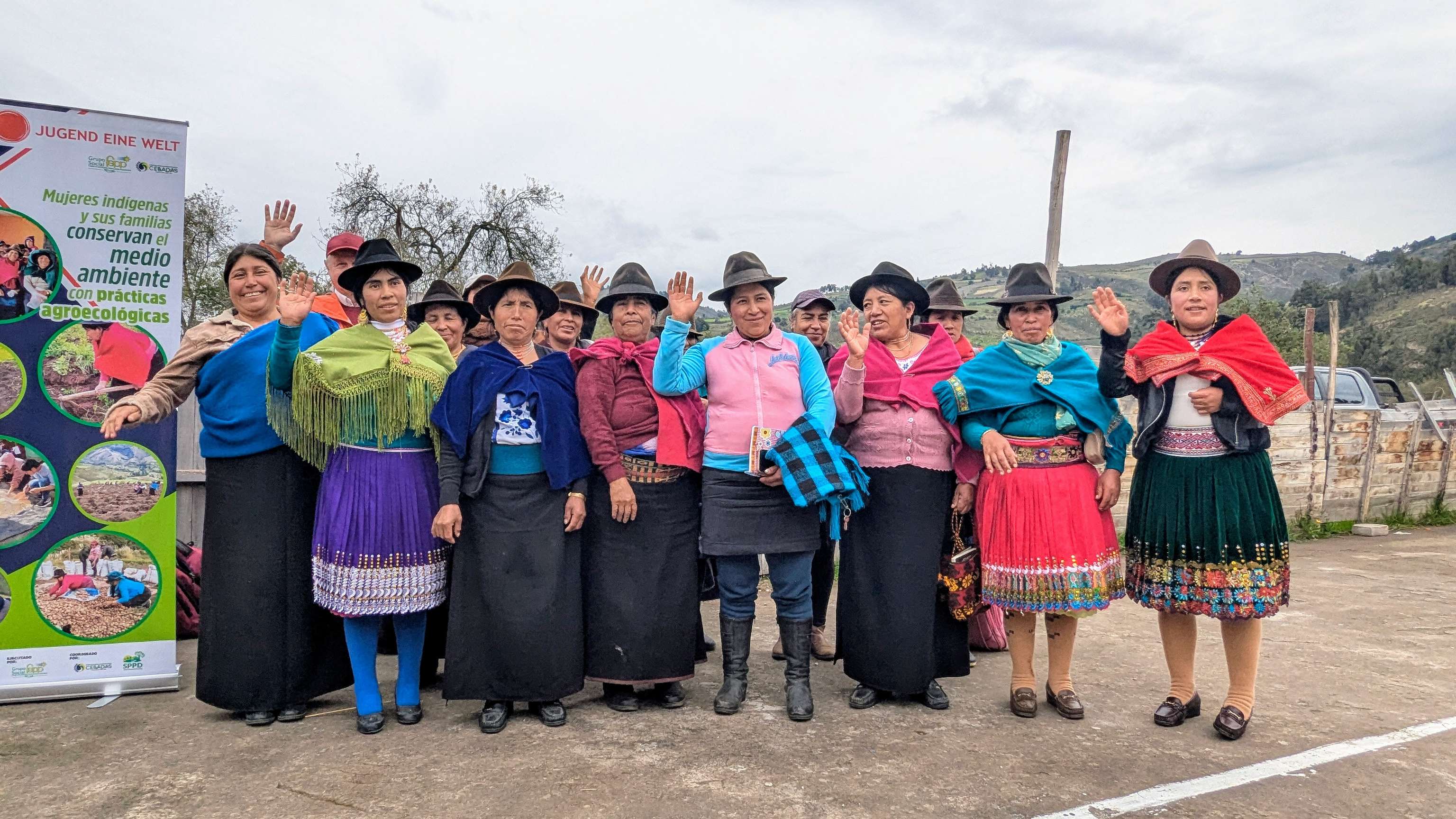 Indigene Frauen in bunter Tracht winken beim Projekt „Jugend Eine Welt" zur Agroökologie in Südamerika.