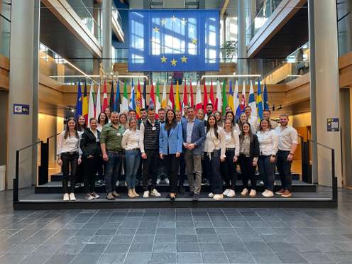 Eine Gruppe von Menschen steht vor den EU-Flaggen in einem modernen Gebäude.