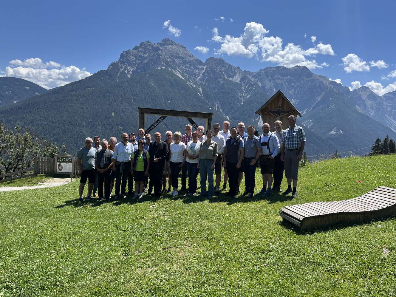 Gruppe von Personen auf einer Almwiese vor beeindruckender Alpenkulisse mit blauem Himmel und einem Holzschild im Hintergrund.