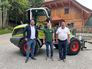 Drei Männer stehen vor einem grünen Traktor auf einem Kiesweg, im Hintergrund ein Holzhaus und Bäume.
