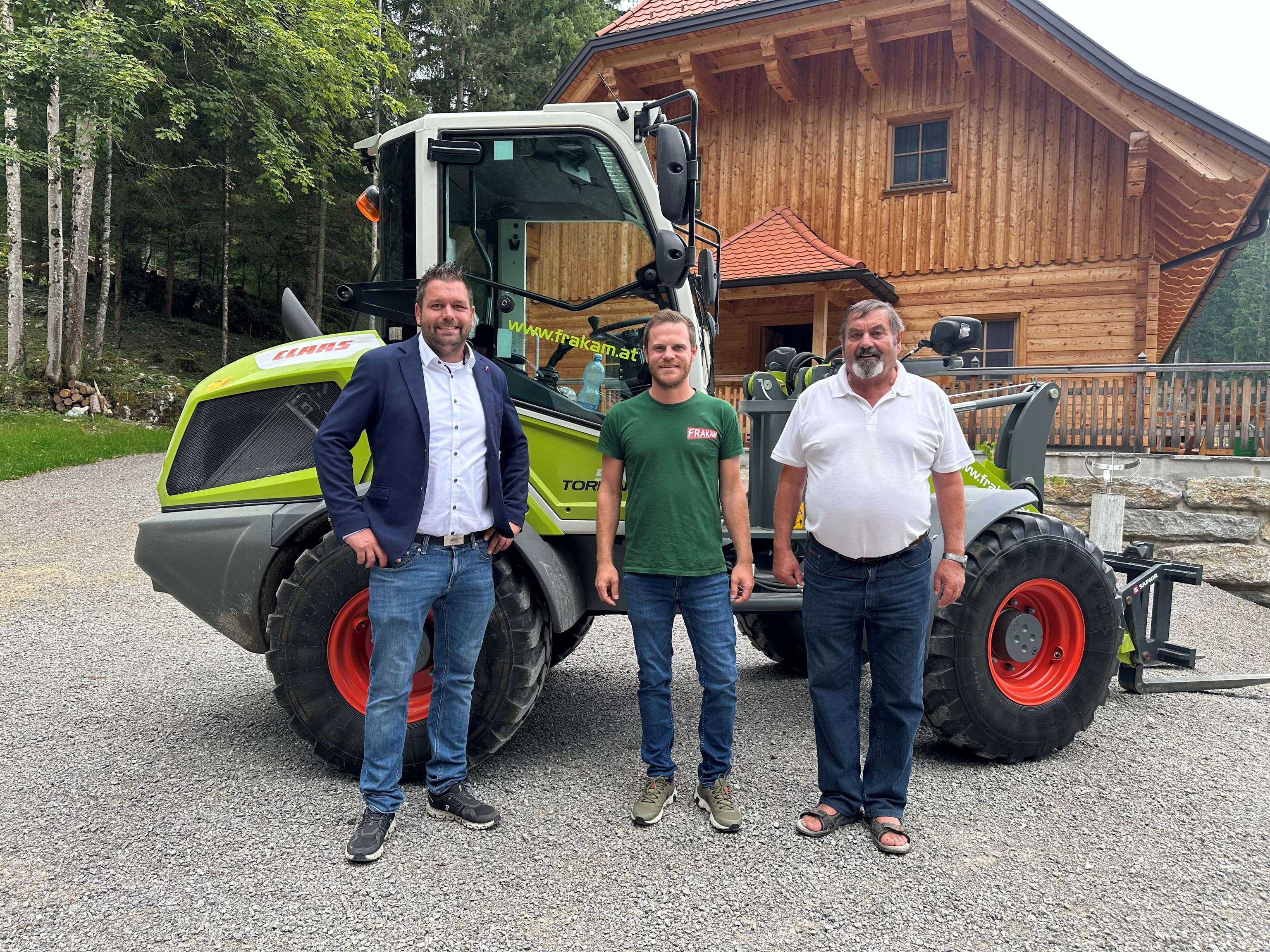 Drei Männer stehen vor einem grünen Traktor auf einem Kiesweg, im Hintergrund ein Holzhaus und Bäume.