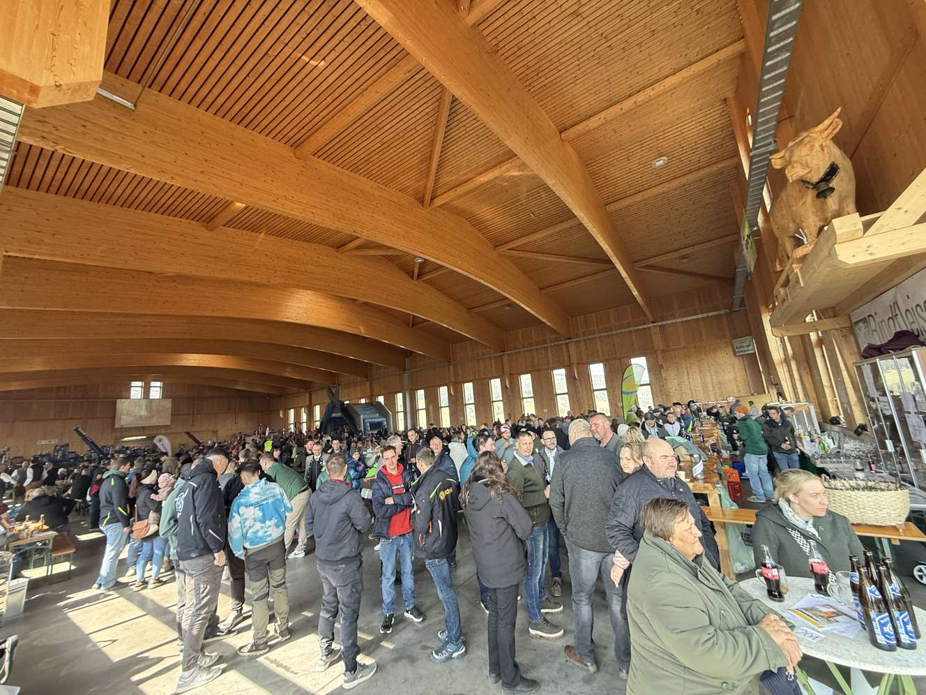 Große Menschenmenge in einer Holzhütte mit gewölbtem Dach, die sich unterhält und Getränke genießt. Helme sind auf Tischen zu sehen.